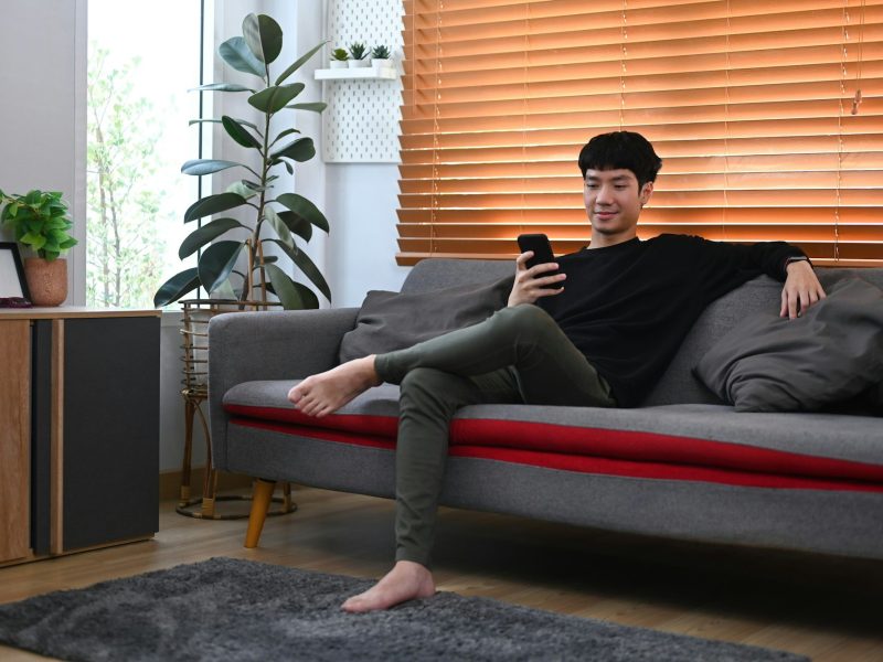 Full length of man using mobile phone on couch at home, spending leisure in cozy winter.